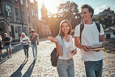Eine junge Frau und ein junger Mann laufen auf einer sonnigen Kopfsteinpflasterstraße. sie halten jeweils Bücher und unterhalten sich. Im Hintergrund ein historisches gebäude und weitere Personen.