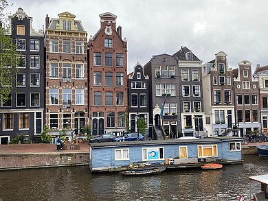 Häuserzeile mit Grachten in Amsterdam. In den Grachten liegt ein blaues Hausboot. 