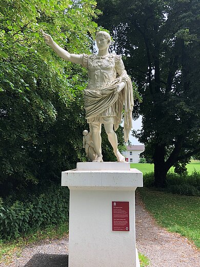Statue von Kaiser Augustus auf einem Sockel im Park. Im Hintergrund befinden sich Bäume und in der Ferne ein Haus.