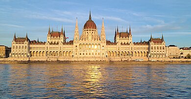 Das ungarische Parlamentsgebäude am Flussufer von der Donau aus fotografiert. Die Sonne spiegelt sich in den Fenstern und im Wasser.