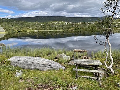 Blauer See in Norwegen umrahmt von grünen Wiesen und einem kleinen Hügel. Auf einer Wiese liegt ein Holzboot. 