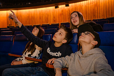 Kinder und Jugendliche sitzen im Planetarium und schauen in den Sternenhimmel in der Planetariumskuppel.

