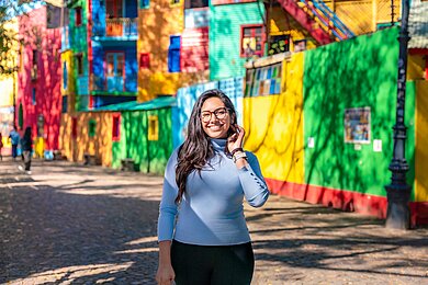 Straßenszene im Viertel Caminito, Buenos Aires, Argentinien. Eine junge Frau mit Brille und blauem Pullover steht vor bunten Häuserfassaden.