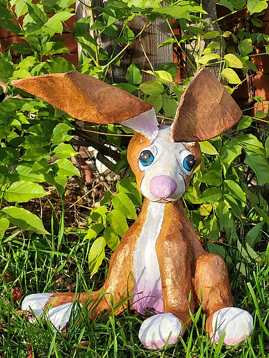 Das Bild zeigt eine handgefertigte Pappmaché-Hasenfigur mit großen braunen Ohren, blauen Augen und rosa Nase. Sie sitzt im Gras vor grünen Blättern, wirkt freundlich und verspielt.