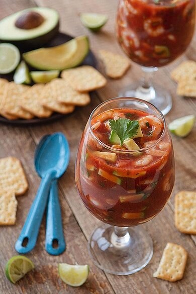 Ein gefülltes Glas in dem ein Shrimp Cocktail abgefüllt ist. Daneben wurde dekoriert mit Avocado, Salsa und Tortillachips.