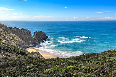 Hinter einer Steilküste in Portugal erstreckt sich weites Meer. Unterhalb der Klippe liegt ein kleiner Strandstreifen.