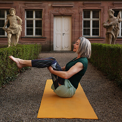 Lächelnde Frau sitzt seitlich auf einer gelben Yogamatte vor einem Gebäude, hat die Beine angewinkelt in Brusthöhe vom Boden abgehoben und umfasst mit beide Hände die Kniekehlen. 