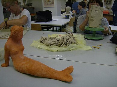 Personen, die in einem Werkraum Ton modellieren, im Vordergrund sieht man eine modellierte Meerjungfrau, daneben und im Hintergrund Köpfe, die gerade modelliert werden.