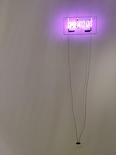Leuchtendes Neonschild mit dem Wort 'heimat' an einer Wand mit zwei langen Kabeln, die zu einer Steckdose führen