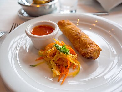CHẢ GIÒ Auf einem weißen Teller ist eine Frühlingsrolle mit einer süß-sauer Soße und einem kleinen Salat garniert.