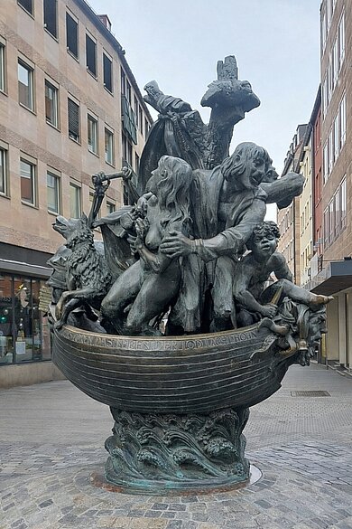 Bronzefigurengruppe mit mehreren menschlichen Figuren und einem Tier auf einem Boot, das auf stilisierten Wellen steht, umgeben von Gebäuden in einer Fußgängerzone