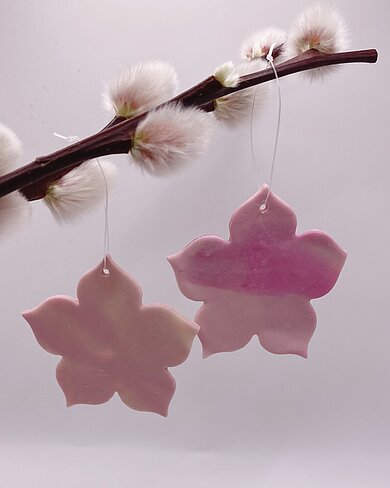 Das Bild zeigt zwei rosafarbene Dekoblumen aus Polymer Clay in Blütenform, die an einem Zweig mit flauschigen Weidenkätzchen hängen.