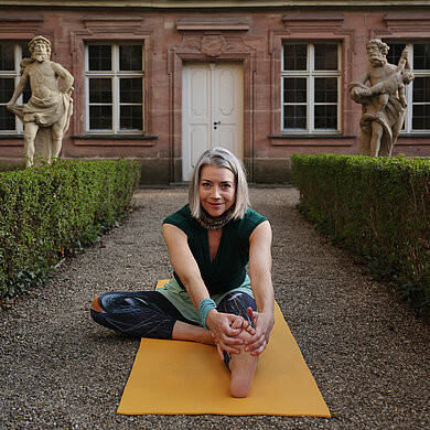 Yoga im Fokus: Yoga- und Beckenbodentraining – Kraft aus der Mitte Eine Frau sitzt auf einer gelben Yogamatte, lächelt in die Kamera und führt eine Yogapose aus.