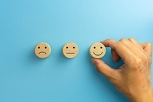Drei Holzscheiben mit Smiley-Gesichtern: unzufrieden, neutral, glücklich, vor blauem Hintergrund.