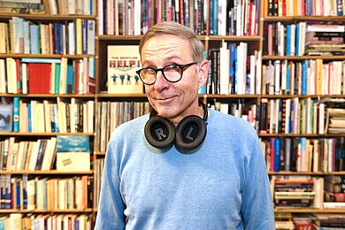 Ein lächelnder Mann im blauen Strickpullover mit Brille und Kopfhörern vor einem Bücherregal