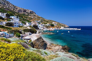 Sicht auf den Hafen der Insel Thimena, Griechenland. Neben einem Strandstück liegen kleine Boote im Wasser, an Land mehrere weiße Häuser und Vegetation.