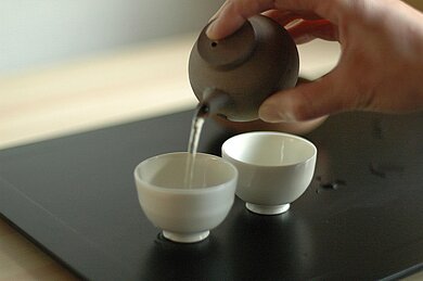 zwei Tassen und eine Teekanne. Eine Tasse wird gerade mit einer klaren Flüssigkeit gefüllt.