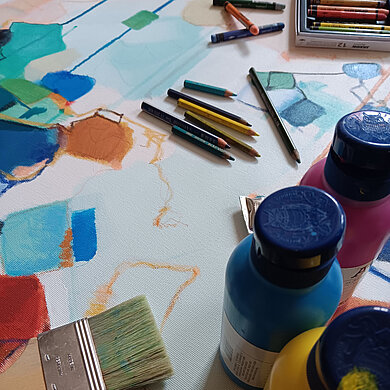 Bild mit einer bunt bemalten Leinwand, im Vordergrund mehrere Farbbehälter, Pinsel und Farbstifte.