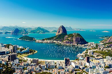 Blick von einem Berg aus auf Rio de Janeiro mit der Skyline vieler Hochhäuser, dem angrenzenden Strand und dem aus dem Meer ragenden Zuckerhut. 