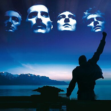 Im Vordergrund die Silhouette von Freddie Mercury, im Hintergrund vor einem blaugefärbtem Sonnenuntergang die Köpfe der 4 Bandmitglieder nebeneinander.
