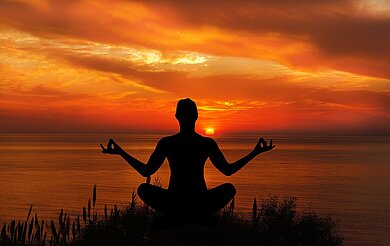 Eine Person sitzt in einer Meditationshaltung draußen in der Natur, in der Abendsonne.