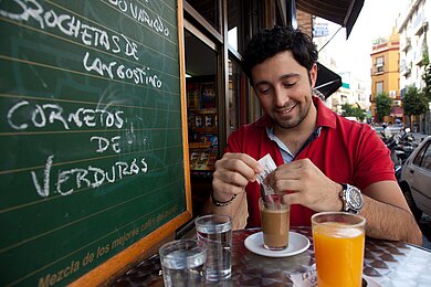 Junger Mann sitzt an einem Tisch im Straßencafé und streut Zucker in seinen Kaffee. Neben ihm steht eine Tafel mit der Speisekarte.