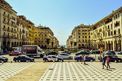 Großer städtischer Platz, der mit hohen Gebäuden umrandet ist. Auf dem Platz parken und fahren Autos; Fußgänger schlendern vorbei.