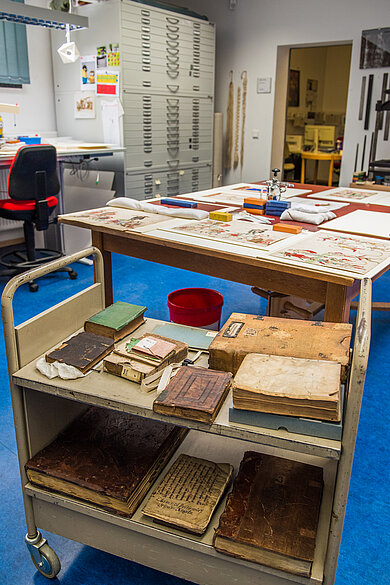Rollwagen mit alten, teils beschädigten Büchern in einem Raum mit Arbeitstisch, Stuhl und Schubladenschrank im Hintergrund.