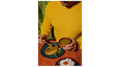 Gorditas jarochas Ein Mann mit gelbem Pullover sitzt beim Essen, hat eine Schale in der Hand und gibt über die Hauptspeise eine Soße.