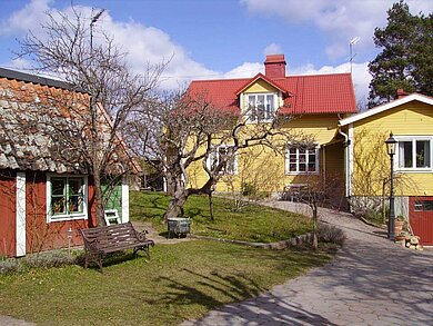 Ein rotes Holzhaus und zwei gelbe Holzhäuser, davor Bäume  und ein Weg, der zu den Häusern führt. Vor dem roten Haus steht eine Bank. 