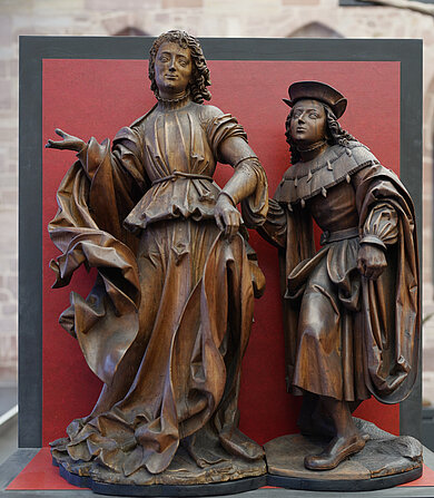 Zwei bronzene Statuen in historischer Kleidung, eine mit ausgestrecktem Arm, die andere mit Umhang und Hut, vor rotem Hintergrund.