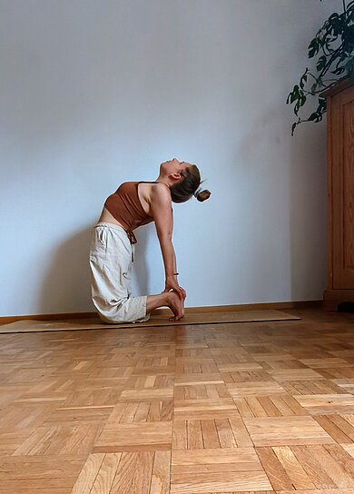 Eine Frau macht eine Yogapose auf einem Holzboden, vor einer weißen Wand.