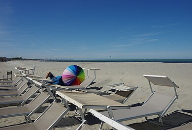 Sandstrand mit buntem Sonnenschirm auf einer Liege. Dahinter das Meer.