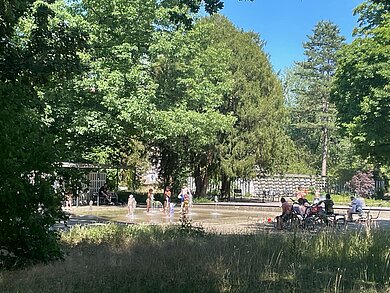 grüner Park, mittig im Bild ein Springbrunnen, in dem Kinder spielen. Es ist Sommer