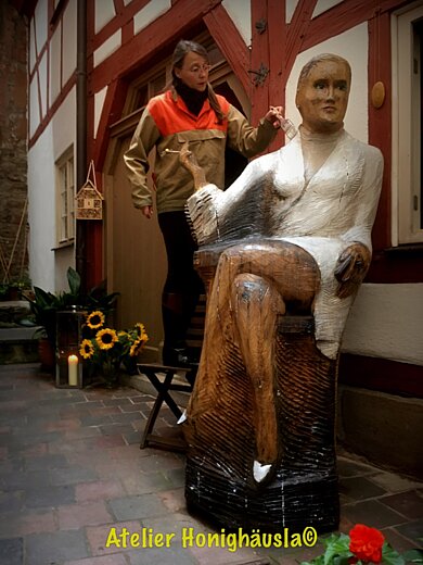 Kunst. Hand. Werk Holzskulptur einer sitzenden Frau mit weißem Oberteil und brauner Hose vor einem Fachwerkhaus, daneben Sonnenblumen und Kerzen.