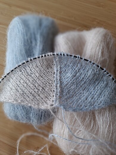 Ein Strickprojekt in zarten Blau- und Rosatönen mit passender Wolle im Hintergrund.