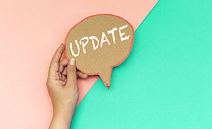Eine Hand hält ein Pappschild mit dem Wort „UPDATE“ vor einem zweigeteilten Hintergrund in Rosa und Türkis.