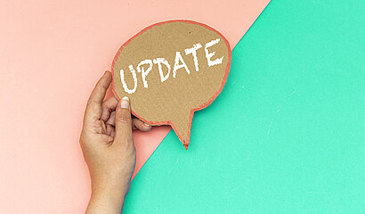 Eine Hand hält ein Pappschild mit dem Wort „UPDATE“ vor einem zweigeteilten Hintergrund in Rosa und Türkis.