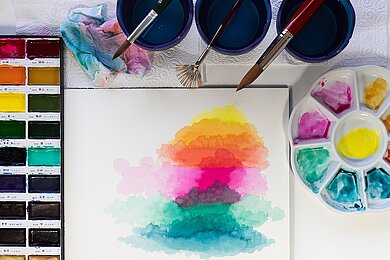 Malkurs: Faszination Aquarell Blick auf einen Aquarellmalkasten und Malpapier mit verlaufenden Aquarellfarben, verschiedene Pinsel sowie eine Mischpalette.
