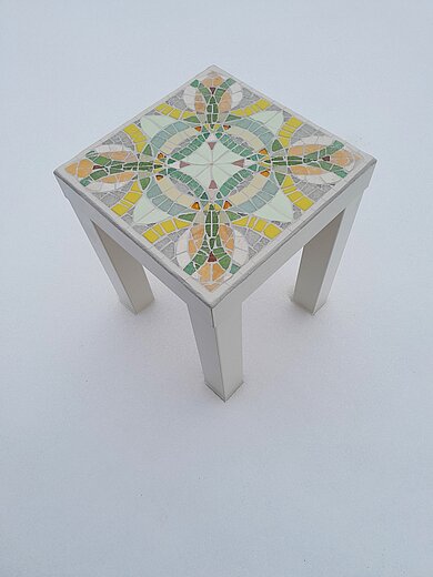 Abgebildet ist ein kleines Tischchen deren Tischplatte mit einem grafischen Mosaikmuster gestaltet wurde in den Farben grün, gelb, weiß, grau, rose und mit roten Steinen.
