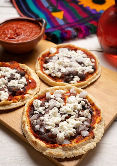Sopes Auf einem Holzbrett liegen drei Ausführungen der mexikanischen Spezialität Sopes. Im Hintergrund ist in einer Schüssel eine Salsa aus Tomaten abgebildet.