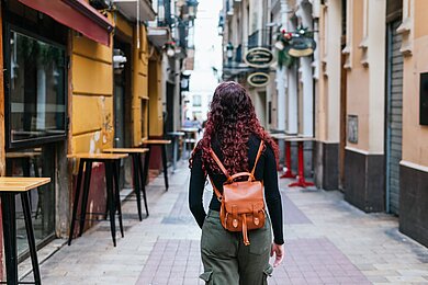 Junge Frau läuft durch eine enge Gasse mit bunten Häuserfassaden und den Stehtischen eines Cafés.