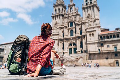 Frau mit Rucksack sitzt am Boden und sieht auf die Fassade einer Kathedrale in Santiago de Compostela.