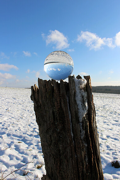 Glaskugel auf einem Baumstumpf in der sich der Himmel und der schneebedeckte Boden spiegeln.