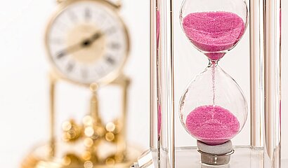Das Bild zeigt eine kleine Standuhr. Daneben eine Sanduhr.