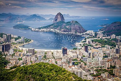 Blick von einem Berg aus auf Rio de Janeiro mit der Skyline vieler Hochhäuser, dem angrenzenden Strand und dem aus dem Meer ragenden Zuckerhut. 