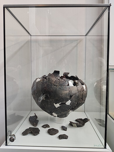 Zerbrochene, zusammengesetzte Keramikvase in einer Glasvitrine mit mehreren Keramikfragmenten auf dem Vitrinenboden