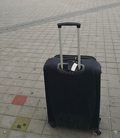 Schwarzer Trolley mit ausgezogenem Griff steht auf gepflastertem Boden mit einzelnen farbigen Steinen