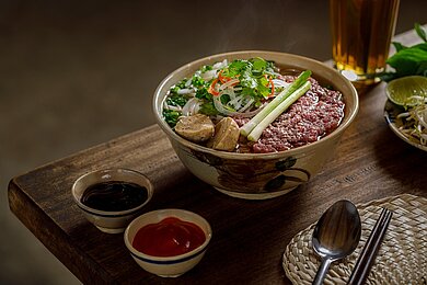 PHỞ BÒ Auf einem Holztisch steht eine große Schüssel gefüllt mit einer traditionellen Pho. Daneben stehen in kleineren Schüsseln Soßen und andere Zutaten, zum Würzen der Pho.