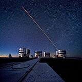 Abgebildet ist das Very Large Telescope (VLT) der Europäischen Südsternwarte unter dem chilenischen Abendhimmel, das gerade einen Laserstrahl zur Anpassung der adaptiven Optik in den Himmel sendet.
